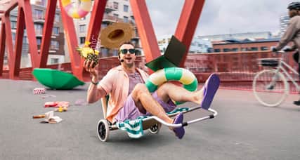 En person i solstol åker över en bro i en stad. Runt omkring flyger strandleksaker, drinkar och parasoll.