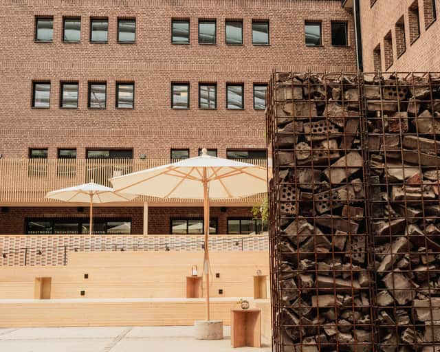 På en atriumgård där man blickar mot en tegelfasad och framför är det ljusa träbänkar och en tegel gabionmur