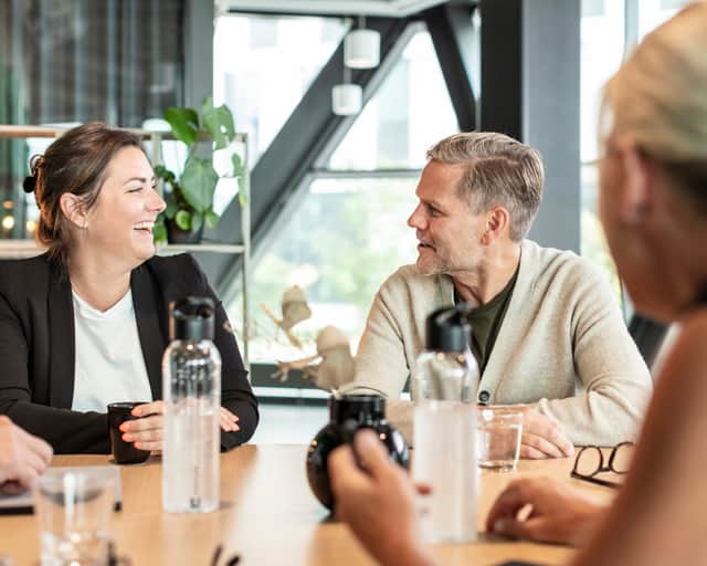 Kvinna och man sitter vid ett bord och tittar på varandra, skrattandes. Fler kollegor sitter runt bordet.