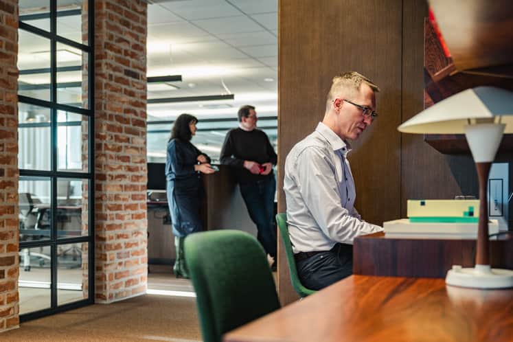 En man arbetar vid sin dator medan två personer pratar i bakgrunden.