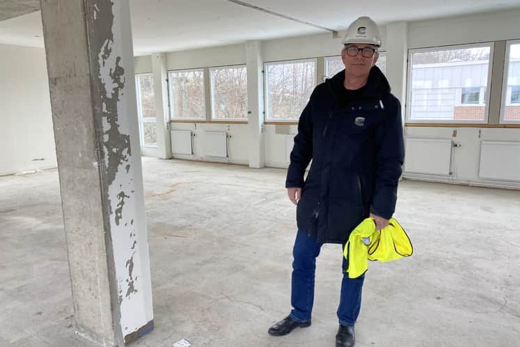 Anders står i en tom lokal med svart jacka, vit hjälp och med en gul byggväst i handen.